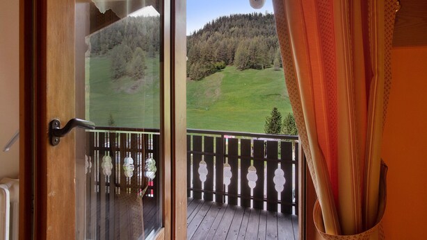 Un balcon en bois avec une porte vitrée donnant sur un champ verdoyant et une colline boisée.