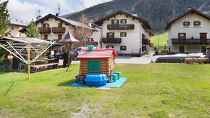 Une cabane en bois avec un toit rouge, une structure de jeu bleue et une zone herbeuse devant.