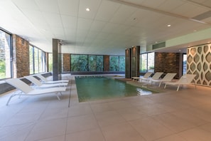 Une piscine intérieure moderne, dotée de chaises longues blanches, de murs en pierre et de grandes fenêtres.