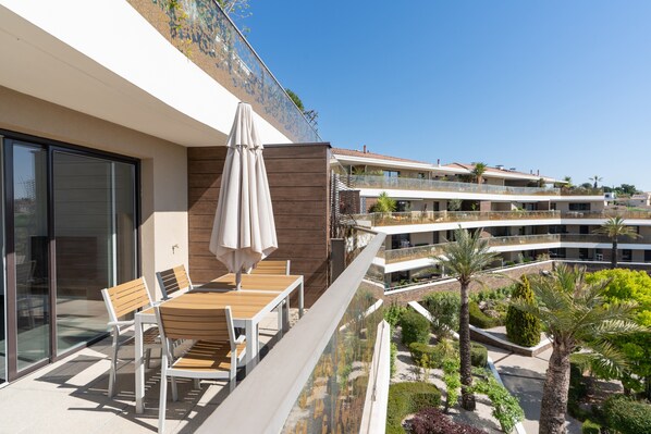 A balcony with a table and chairs, a glass railing, and a view of other buildings and greenery.