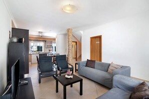 A living room with a sofa, chairs, a coffee table, a TV, and a kitchen in the background.