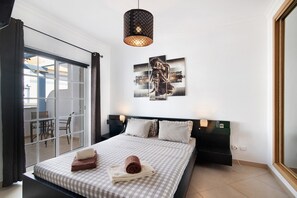 A hotel room with a bed, a large window with curtains, a wall-mounted artwork, and a ceiling light fixture.