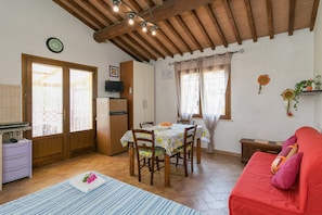 Ein gemütliches Zimmer mit Holzbalkendecke, einem Esstisch mit Stühlen, einem roten Sofa und einer Küchenzeile mit Kühlschrank.