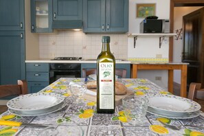 A kitchen with blue cabinets, a table with a lemon-patterned tablecloth, and a bottle of OLIO DI OLIVA on the table.
