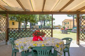 Een overdekt terras met een tafel voor twee personen, omringd door groene stoelen, onder een houten pergola.