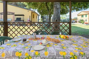 Ein Essbereich im Freien mit einem Tisch für zwei Personen, einem Grill und Blick auf ein gelbes Haus und Bäume.
