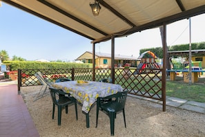 Eine Außenpatio mit Tisch und Stühlen, ein überdachter Bereich und ein Spielplatz im Hintergrund.