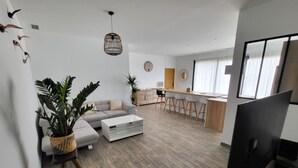 A modern living room with a sofa, coffee table, and a TV. There is a kitchen area with a bar and stools. - La Zenitude, a beautiful single-storey property with well-appointed features (Sérignan)