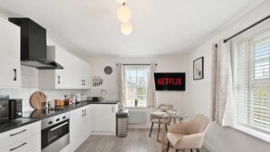 A modern kitchen with white cabinets, a black range hood, and a dining area with a table and chairs. - Tropical Inspired 2-Bedroom Flat (Dunmow)