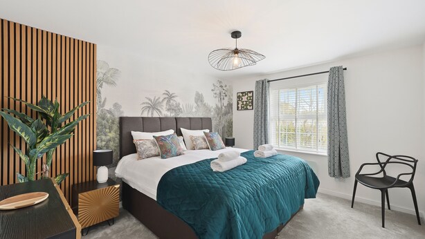 A bedroom with a bed, bedside table, lamp, and a large window with curtains. - Tropical Inspired 2-Bedroom Flat (Dunmow)