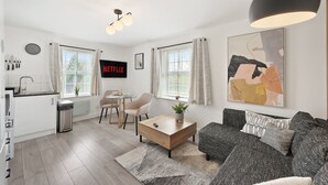 A modern living room with a sofa, coffee table, and artwork. There is a kitchen area with a sink and cabinets. - Tropical Inspired 2-Bedroom Flat (Dunmow)