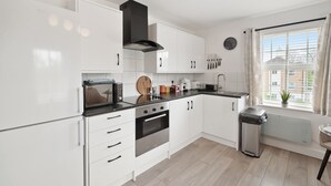 A modern kitchen with white cabinets, a black range hood, a built-in oven, and a window with curtains. - Tropical Inspired 2-Bedroom Flat (Dunmow)