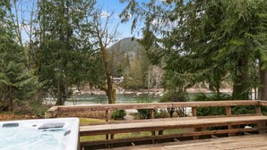 A wooden deck with a hot tub overlooking a river and forested mountains.