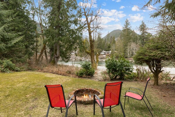A fire pit with two red chairs in a grassy area with a lake and forest in the background.