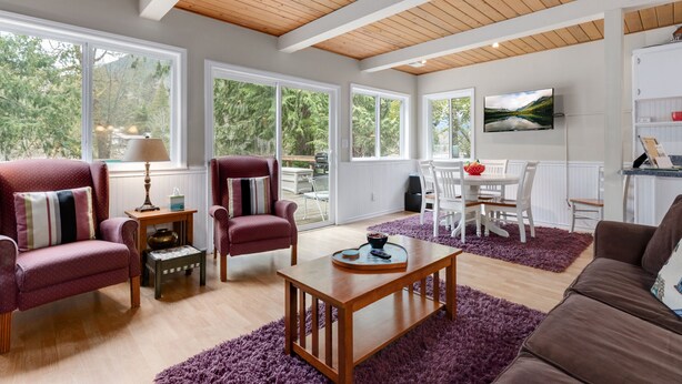 A spacious living room with wooden ceiling beams, large windows, and a view of trees.