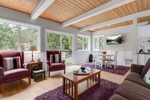 A spacious living room with wooden ceiling beams, large windows, and a view of trees.
