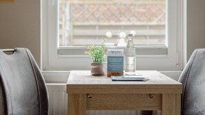 Una pequeña mesa de madera con una planta en maceta, una botella de agua y un folleto informativo en una habitación con vistas a un muro de ladrillo y una valla a través de una ventana.