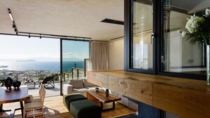 Un salon moderne avec un plancher en bois, un canapé, une table basse et un coin repas avec des chaises. Une grande fenêtre offre une vue sur la ville et les montagnes.
