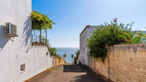 Rua estreita ladeada por edifícios brancos, com vista para o mar ao longe.