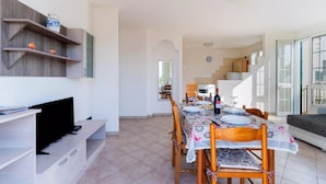 Uma ampla sala de jantar com uma mesa comprida posta para quatro pessoas, cadeiras de madeira e uma televisão sobre um suporte.