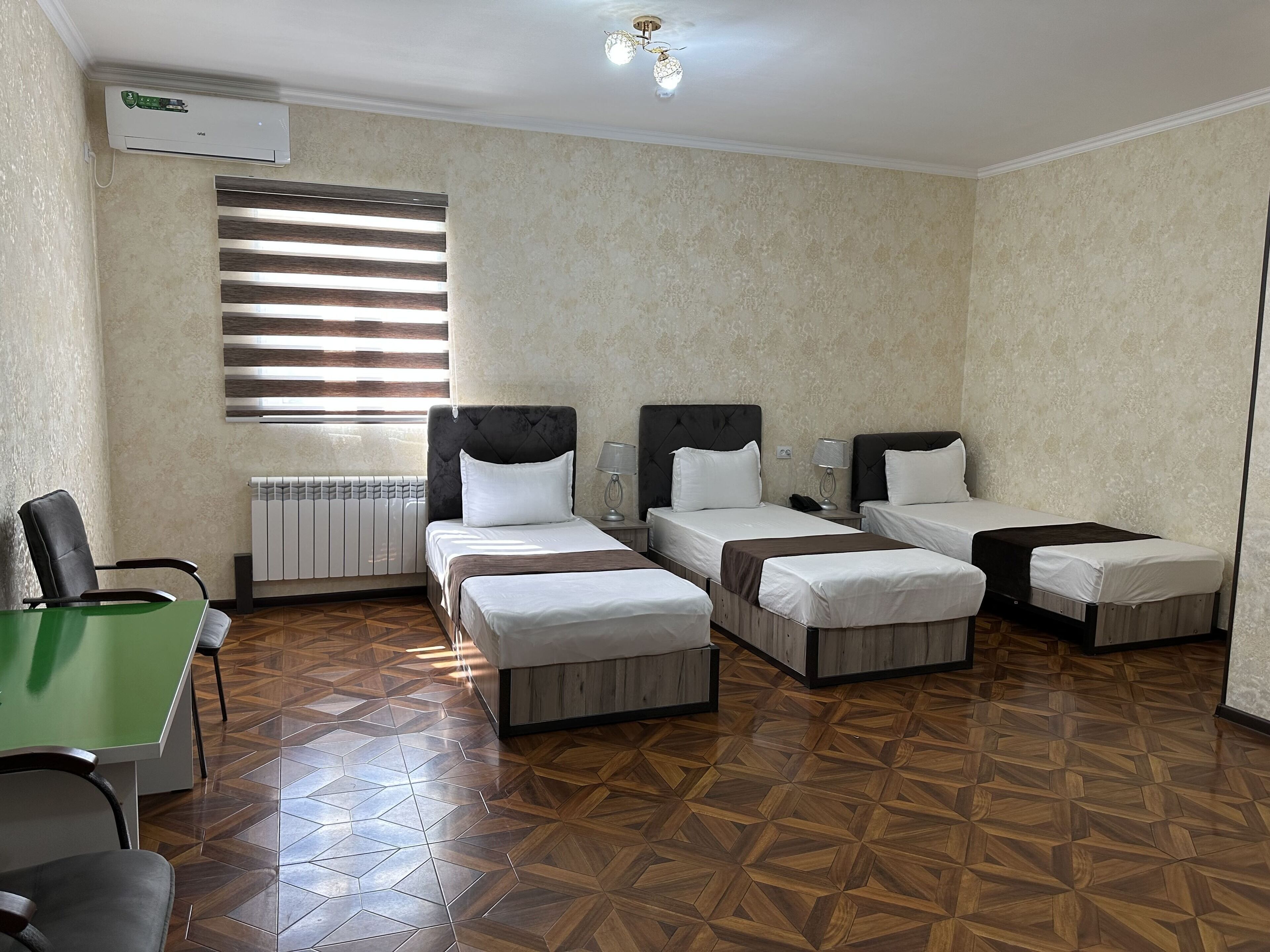 A hotel room with three single beds, a green desk, a chair, a radiator, and a window with blinds.