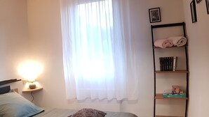 Un dormitorio con cama, una lámpara de noche, una ventana con cortinas translúcidas, un estante con libros y una toalla doblada, y un cuadro colgado en la pared.