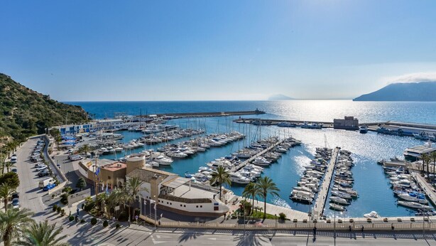 A marina with numerous boats docked, a clear blue sky, and a mountainous backdrop.