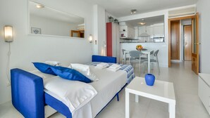 A modern hotel room with a bed, a small table, and a kitchenette in the background.