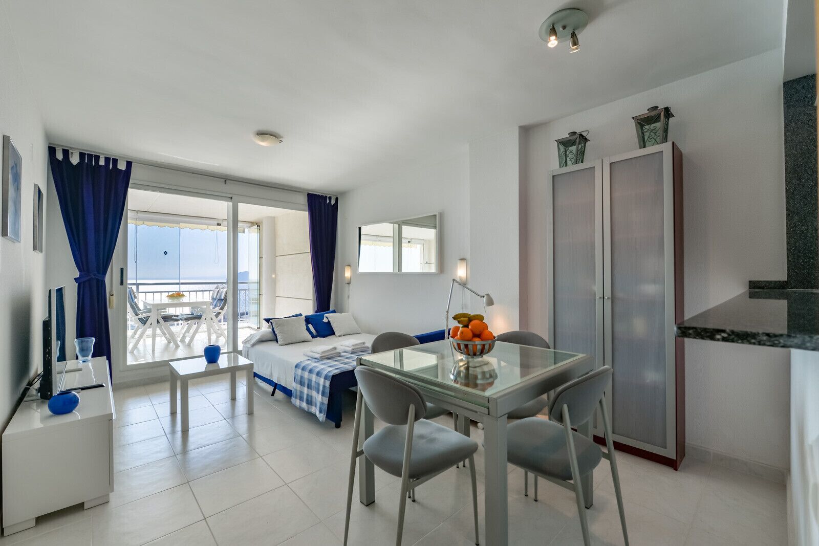 Une chambre d’hôtel moderne avec un balcon, un coin repas comprenant une table et des chaises, un lit recouvert d’une courtepointe à carreaux bleus et blancs, et une armoire.