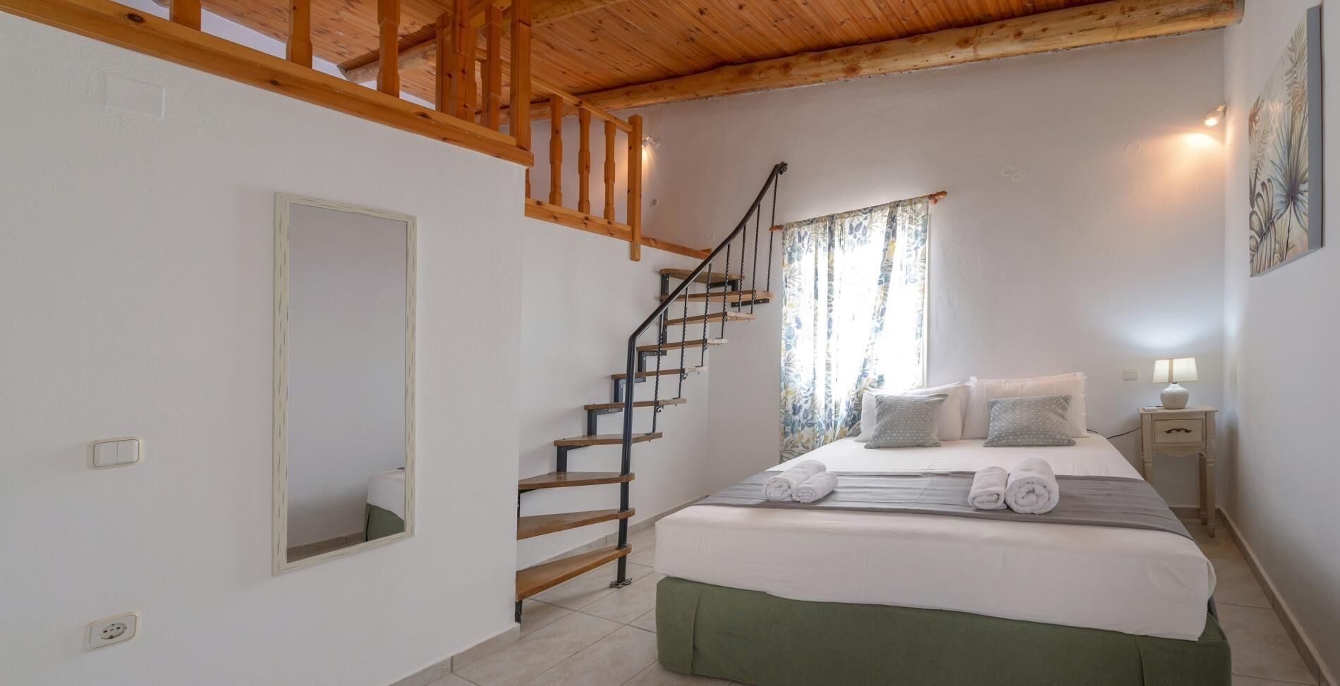 A bedroom with a wooden staircase, a bed with pillows and towels, a bedside table with a lamp, and a window with curtains.