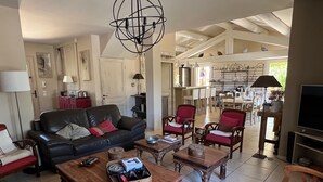 A spacious living room with a black leather sofa, wooden coffee tables, and red armchairs. A chandelier hangs from the ceiling, and a television is mounted on the wall. - Villa de Charme, à Proximité Immédiate D'uzes et du Pont du Gard (Argilliers)