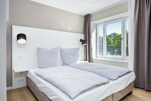 Quarto moderno com uma cama grande, mesa de cabeceira, luminária na parede e janela com cortinas.