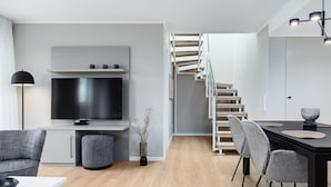 Ein modernes Wohnzimmer mit Flachbildfernseher, grauen Sofas, einem Esstisch mit Stühlen und einer Treppe, die in eine andere Ebene führt.