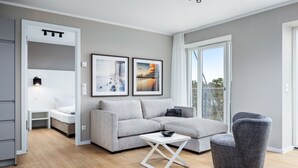 Ein modernes Wohnzimmer mit einem grauen Ecksofa, einem grauen Sessel und einem weißen Couchtisch. An der Wand befinden sich zwei gerahmte Bilder, ein kleiner Beistelltisch und eine eingebaute Regaleinrichtung.