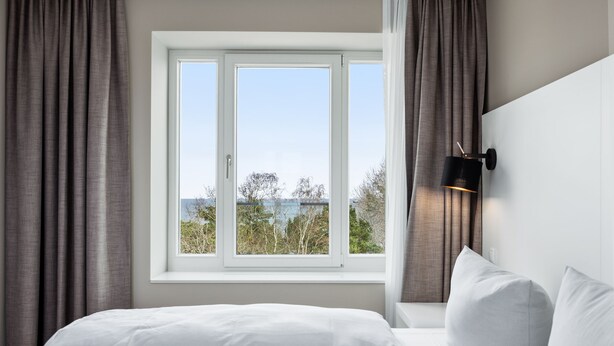Ein Schlafzimmer mit großem Fenster, weißer Bettwäsche und einer Wandleuchte.