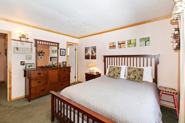 Een slaapkamer met een houten bed, een dressoir en een nachtkastje. Er zijn decoraties aan de muur en een deur naar een andere kamer.