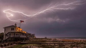 A beachfront house with a balcony overlooking the ocean, a flag on top, and a stormy sky with lightning. - Beachfront, Pool, Spa, Pet Friendly, Elevator, Decks, fire pit, Games! (Saint George Island)