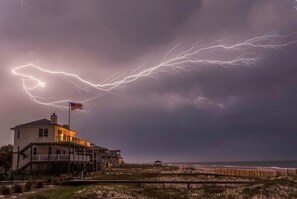 A beachfront house with a balcony overlooking the ocean, a flag on top, and a stormy sky with lightning.