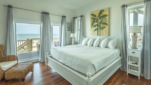 A bedroom with a large bed, a view of the ocean, a chair, and a nightstand with a lamp. - Beachfront, Pool, Spa, Pet Friendly, Elevator, Decks, fire pit, Games! (Saint George Island)