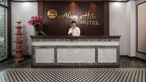 A hotel reception desk with a marble counter, a decorative vase with pink flowers, and a wooden backdrop with the hotel's name. - Nam Hai Hotel (Hanoi)
