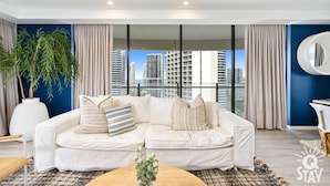 Uma sala de estar moderna com um sofá branco, uma janela grande com vista para a cidade e uma planta em vaso.