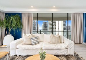 A modern living room with a white sofa, a large window with a city view, and a potted plant. - The Wave - 3 Bedroom Ocean View Unit (Broadbeach)