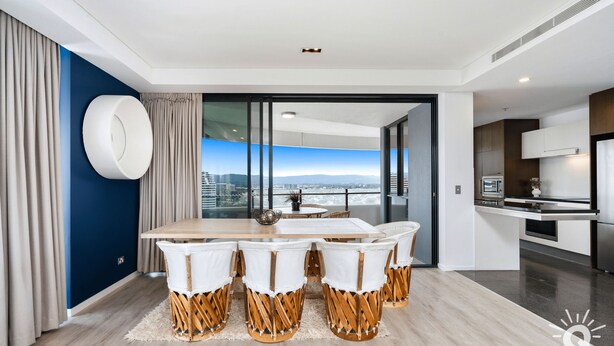 A modern dining area with a wooden table and white chairs, a view of the cityscape through large windows, and a kitchen area with modern appliances. - The Wave - 3 Bedroom Broadbeach Apartment (Broadbeach)