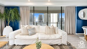 A modern living room with a white sofa, large windows, and a view of high-rise buildings. - The Wave - 3 Bedroom Broadbeach Apartment (Broadbeach)