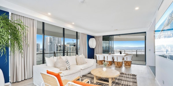 Uma sala de estar moderna com sofá, poltronas, mesa de centro e vista para a paisagem urbana através de grandes janelas.