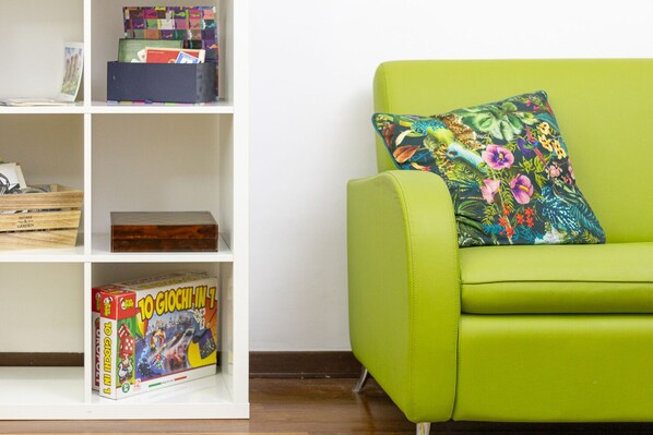 A green sofa with a floral pillow, a white bookshelf with various items including a game box, and a wooden crate labeled "HOME & GARDEN".