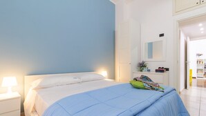 Ein Schlafzimmer mit Bett, Nachttischen, einem Schminktisch und einem Spiegel, sowie einer Tür zu einem anderen Raum.