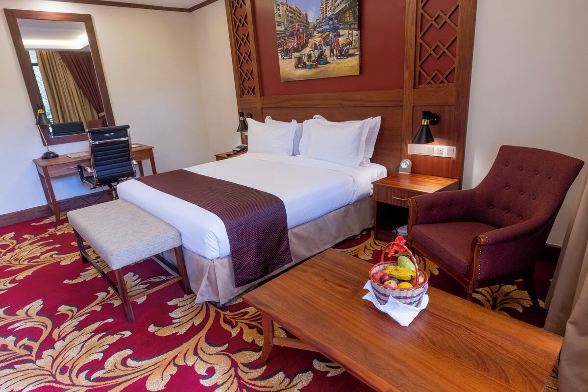 Une chambre d’hôtel avec un lit, un bureau, une chaise, une table avec des fruits, un banc, un miroir et un tableau au mur.