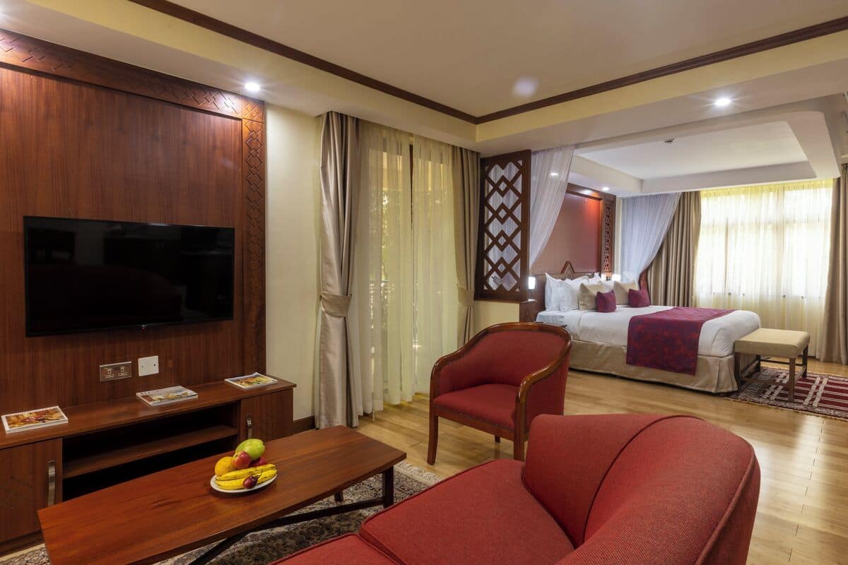 Une chambre d’hôtel avec une télévision, un lit, un fauteuil rouge, une table basse en bois avec des fruits et une table à manger.