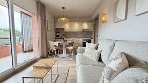 A modern living room with a sofa, a dining table with chairs, a kitchen area, and large windows.